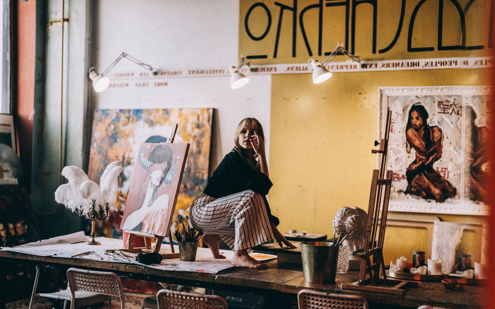 Un’artista seduta sul tavolo del suo studio tra le tele